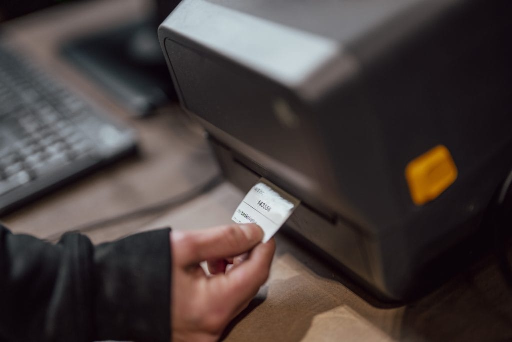 En etiketteskriver printer ut en lapp med nr på seg. En hånd tar i mot lappen.