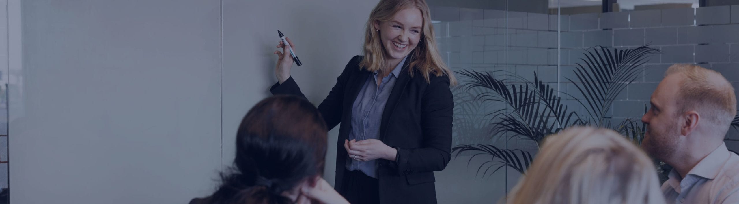 En smilende dame i forretningsklær tegner på et whiteboard foran kollegaene sine og viser nye moduler til styringssystemet Zigma ERP. Foto.
