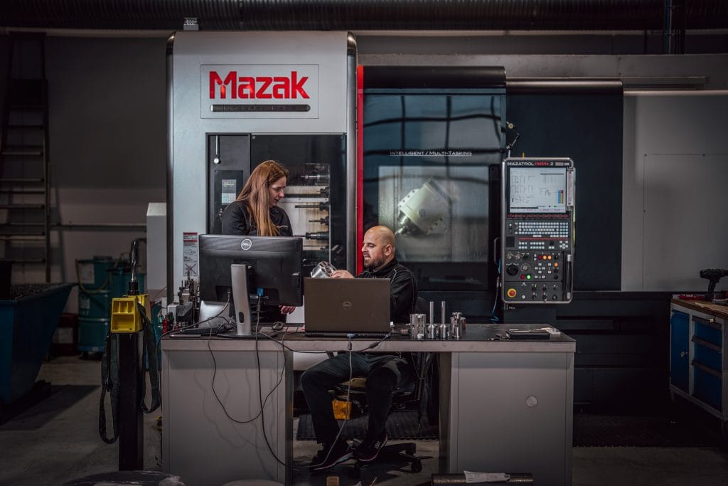 To mennesker i arbeidsklær står foran en mazak-maskin og bak en pc. De har en diskusjon.